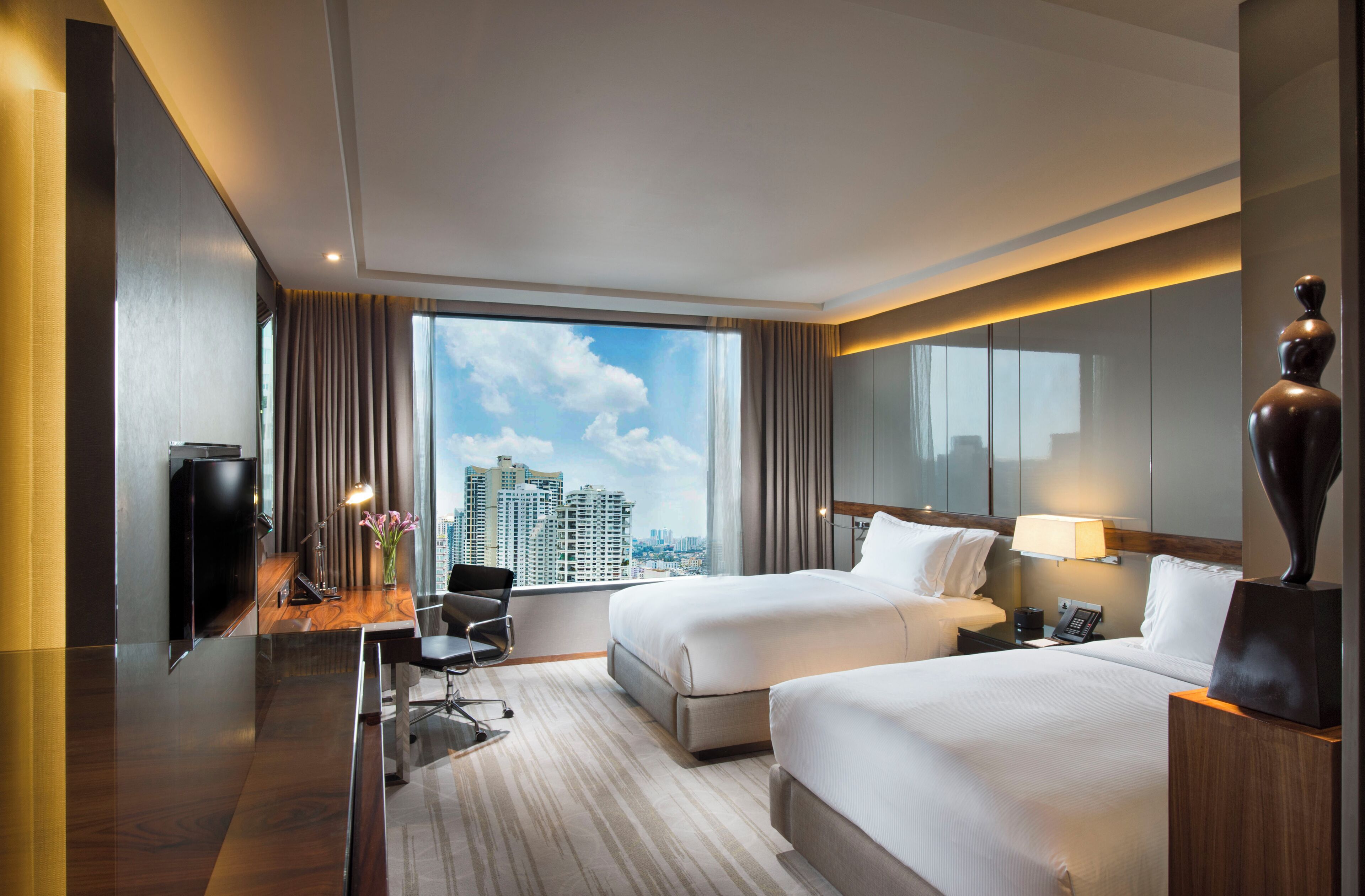 A hotel room with two beds, a desk with a chair, a desk lamp, a telephone, and a large window with a city view.