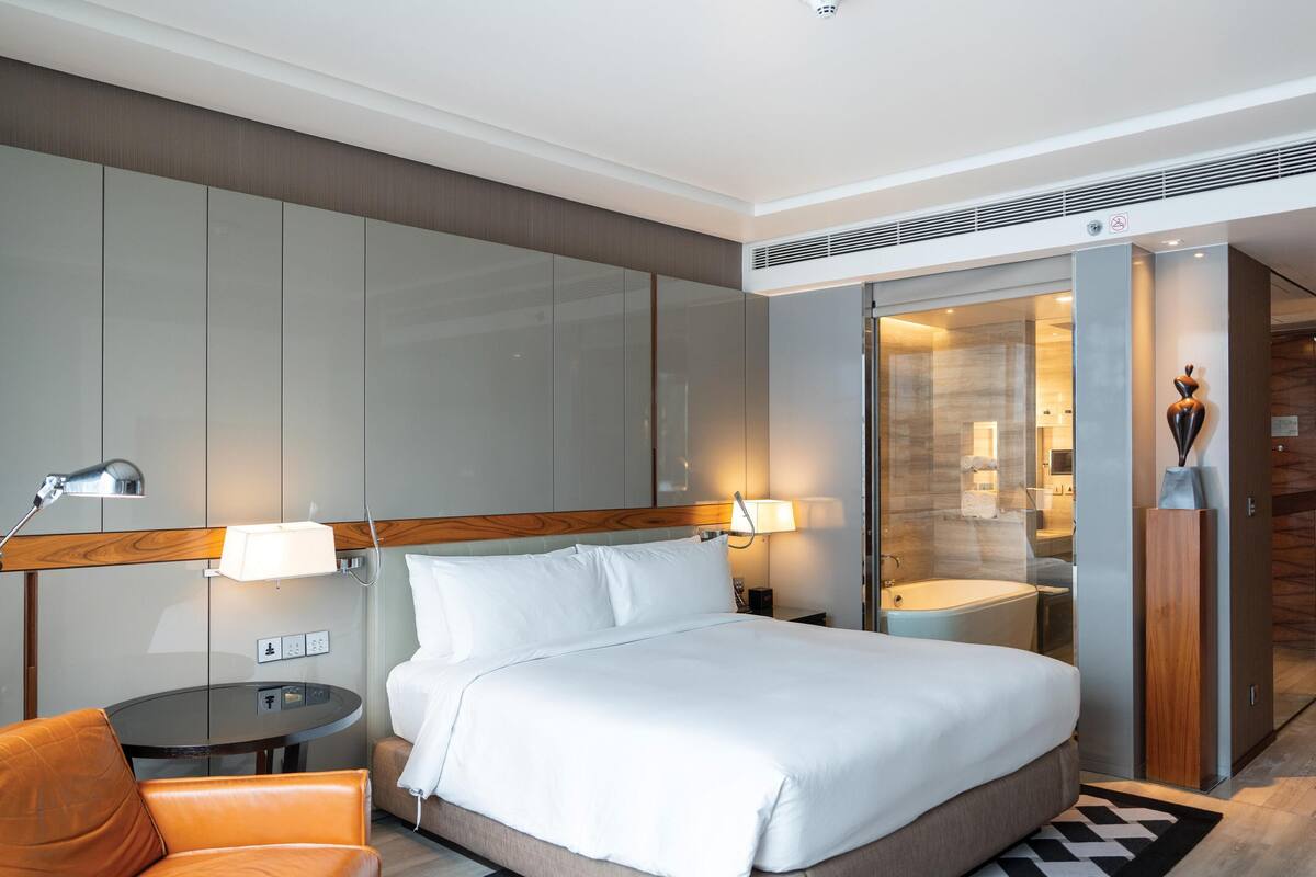 Une chambre d’hôtel moderne équipée d’un lit, d’une chaise, d’un bureau, d’une lampe, d’un miroir et d’une salle de bain visible à travers une porte en verre.