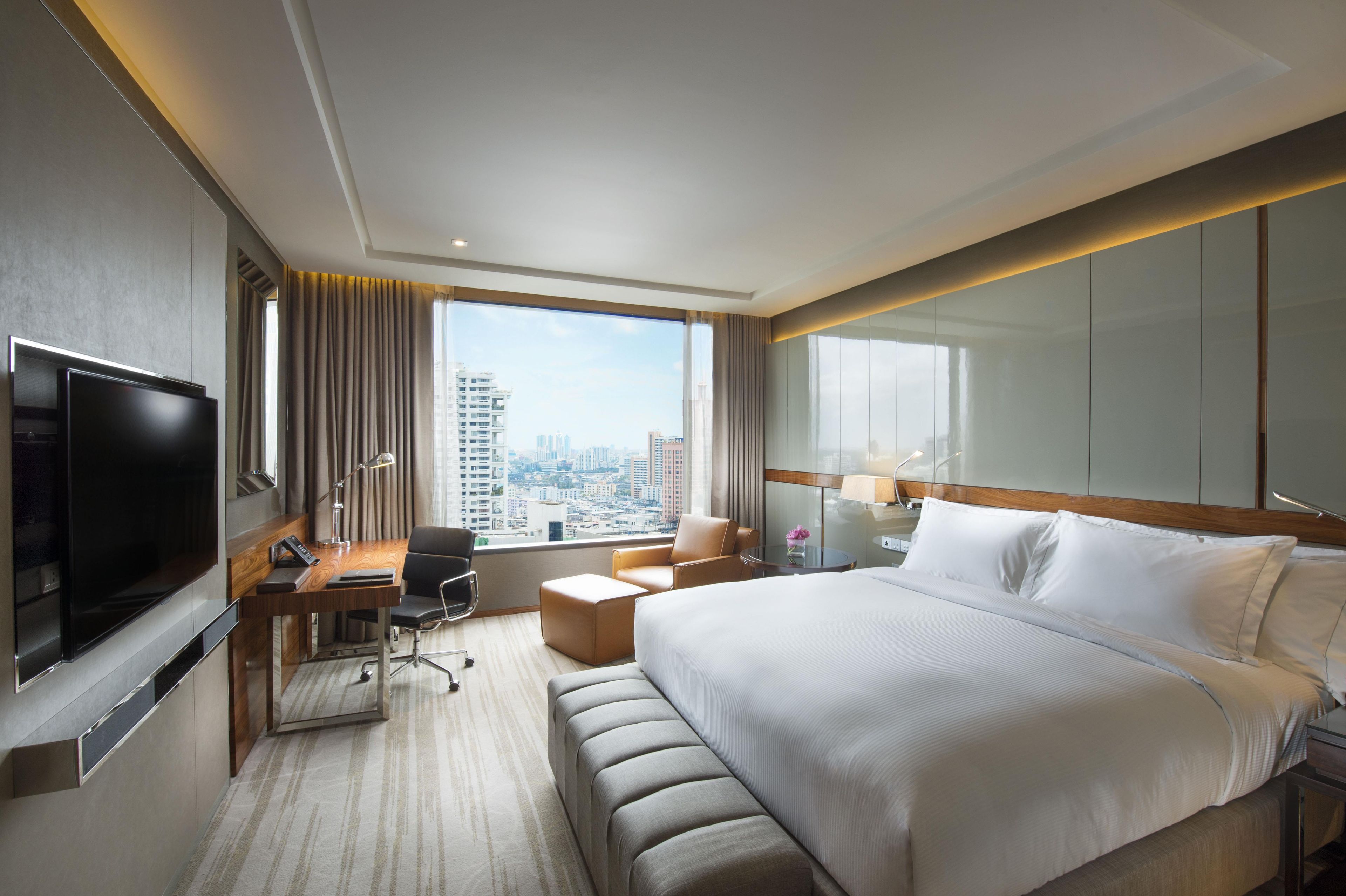 A hotel room with a large bed, a desk with a flat-screen TV, a chair, and a view of the cityscape.