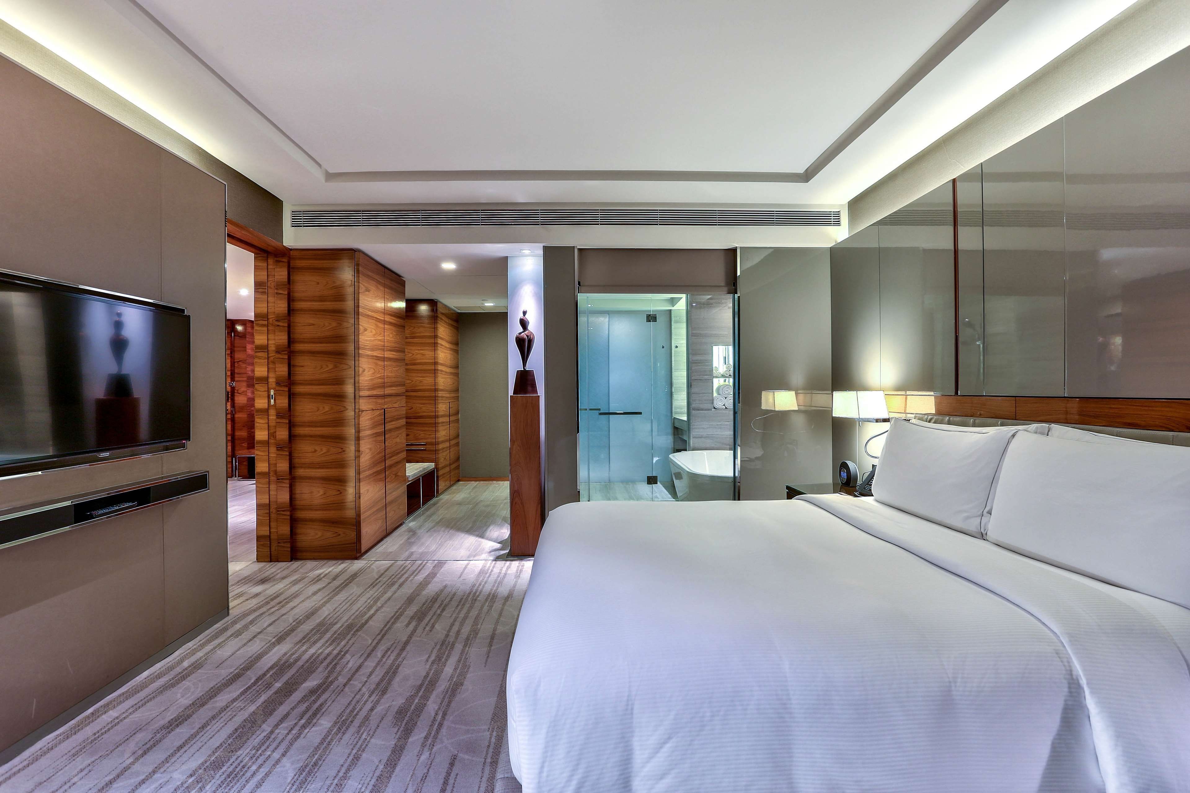 A hotel room with a large bed, a television, a wooden wardrobe, a bathroom, and a mirror.