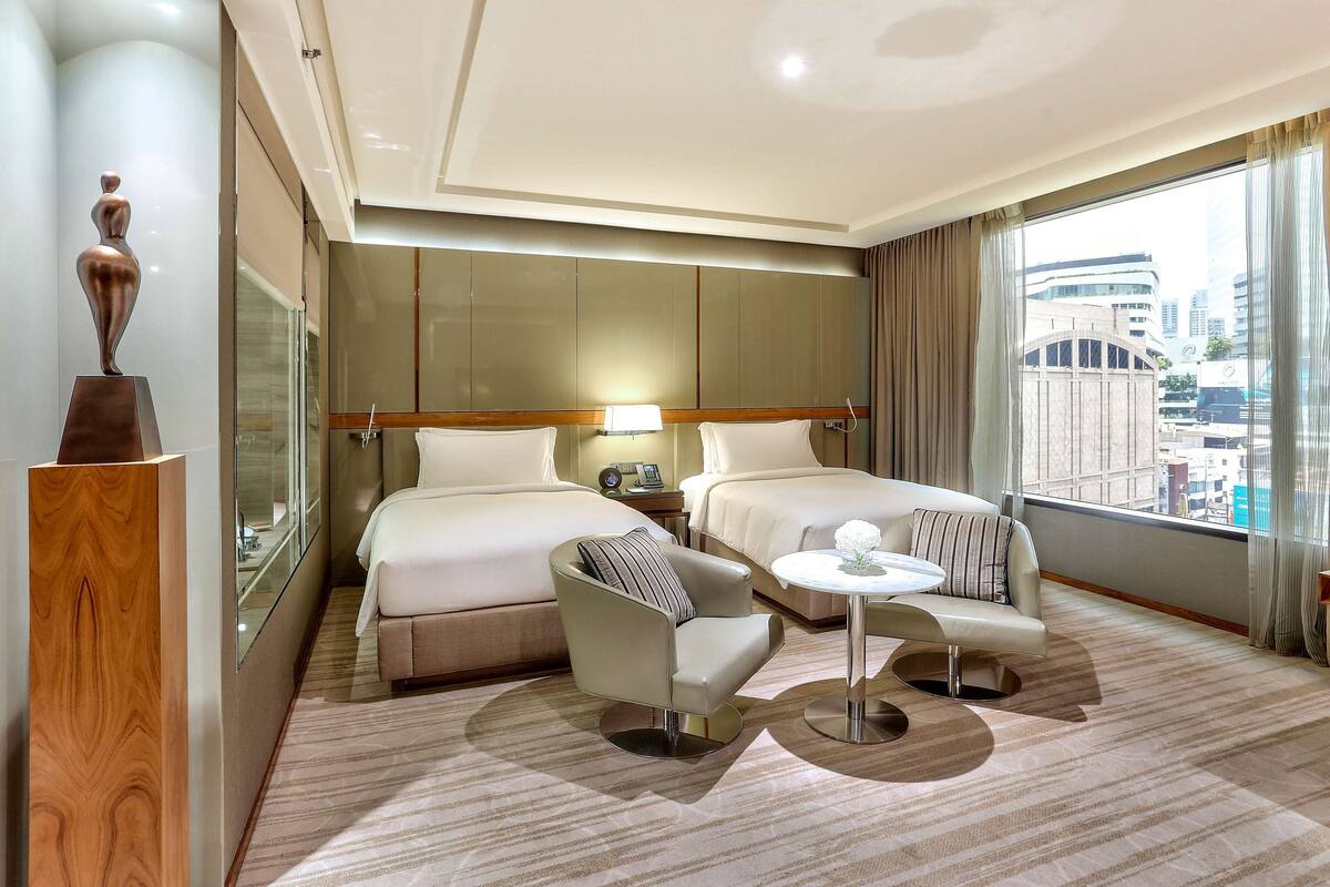 Une chambre d’hôtel avec deux lits, une petite table ronde, une chaise, une table de chevet avec une lampe, et une vue sur la ville grâce à de grandes fenêtres.