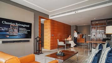 A modern hotel lobby with a television displaying a dessert advertisement, a seating area with leather sofas, a wooden accent wall, and a reception desk.