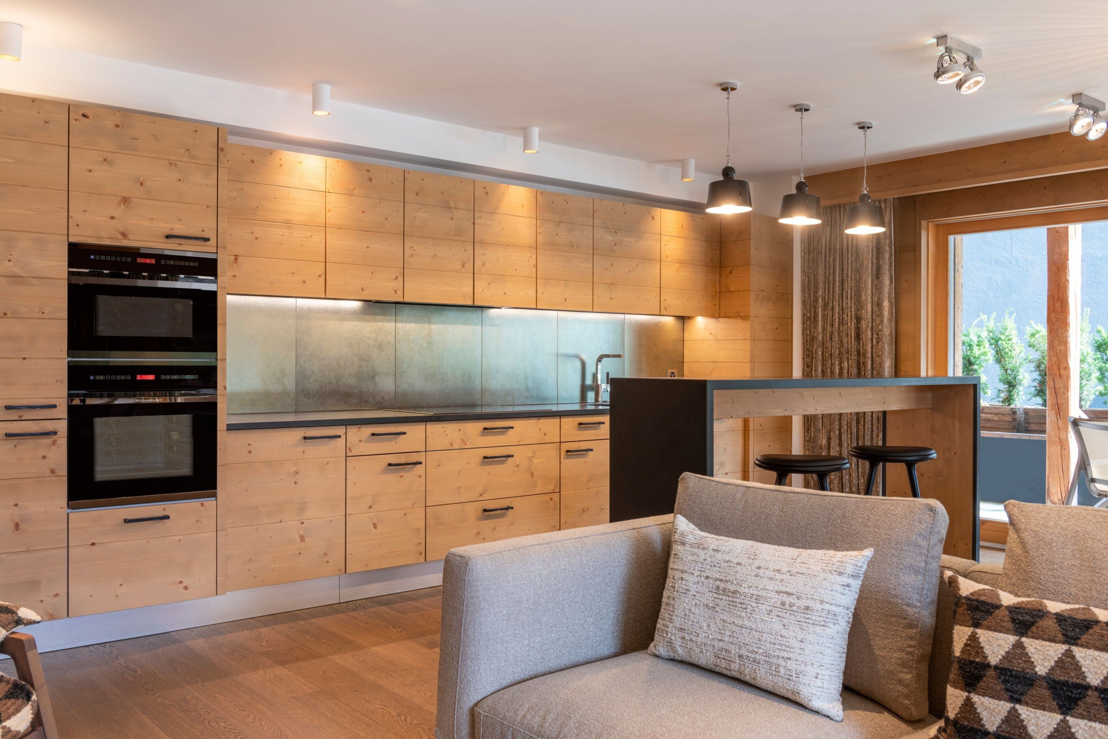 A modern kitchen with wooden cabinetry, built-in ovens, and a dining area with a sofa and bar stools.