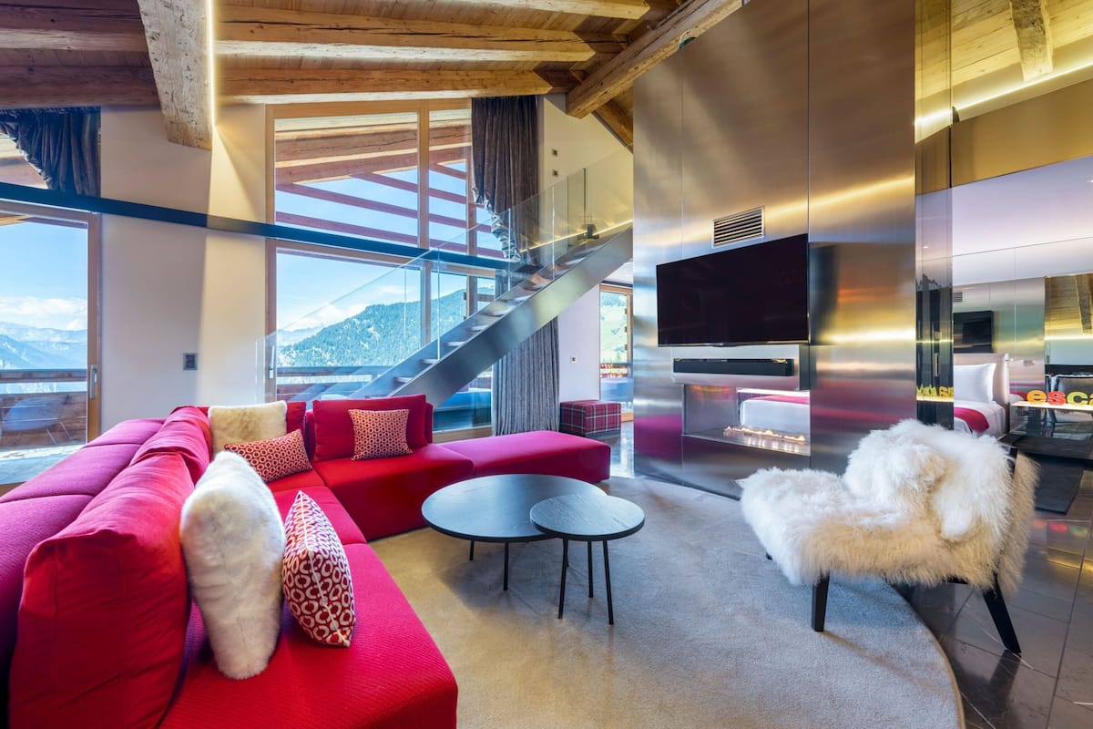Una sala moderna con un sofá rojo, dos mesas redondas y un sillón blanco y mullido. Un televisor grande está montado en la pared, y se puede ver una escalera al fondo.