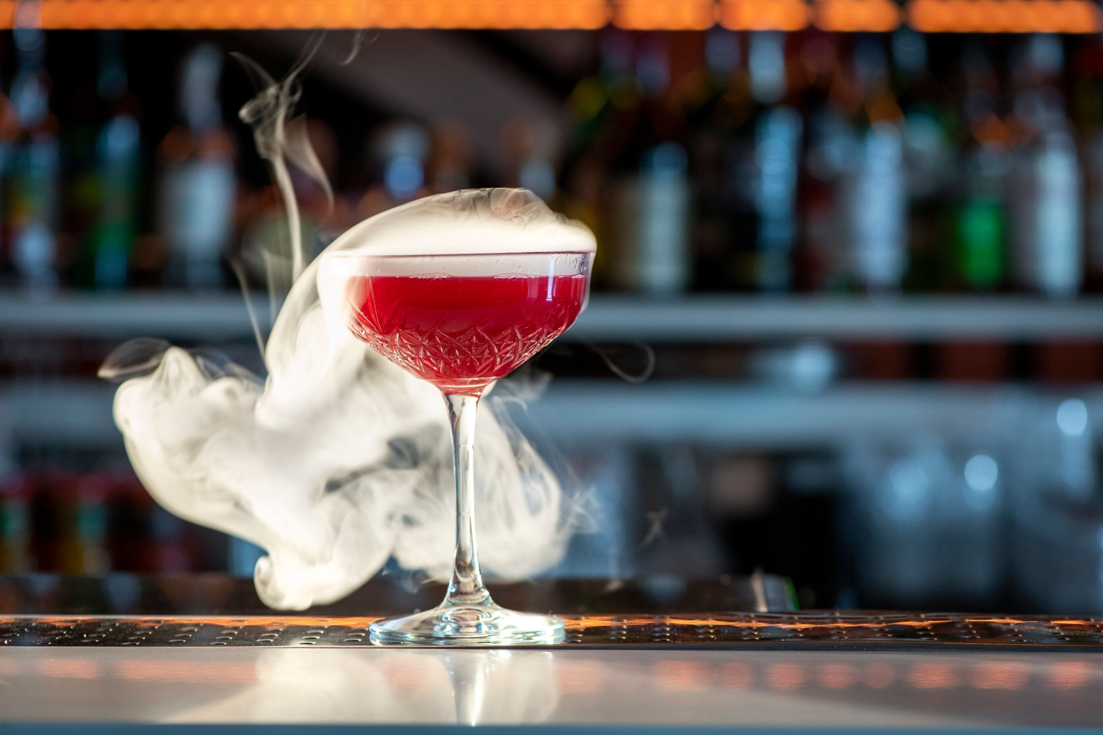 A cocktail in a glass with a foamy top, placed on a bar counter with a blurred background of bottles.