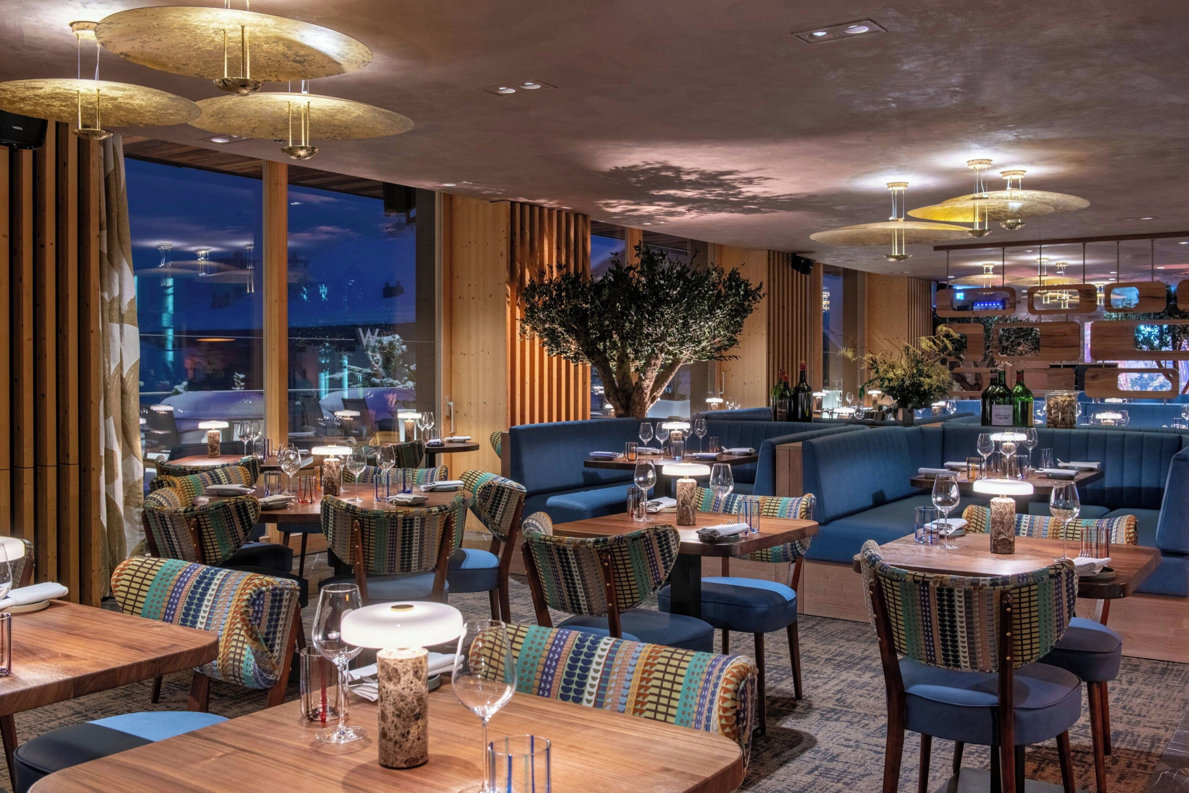A modern restaurant with wooden furniture, blue cushioned seating, and large windows overlooking a cityscape.