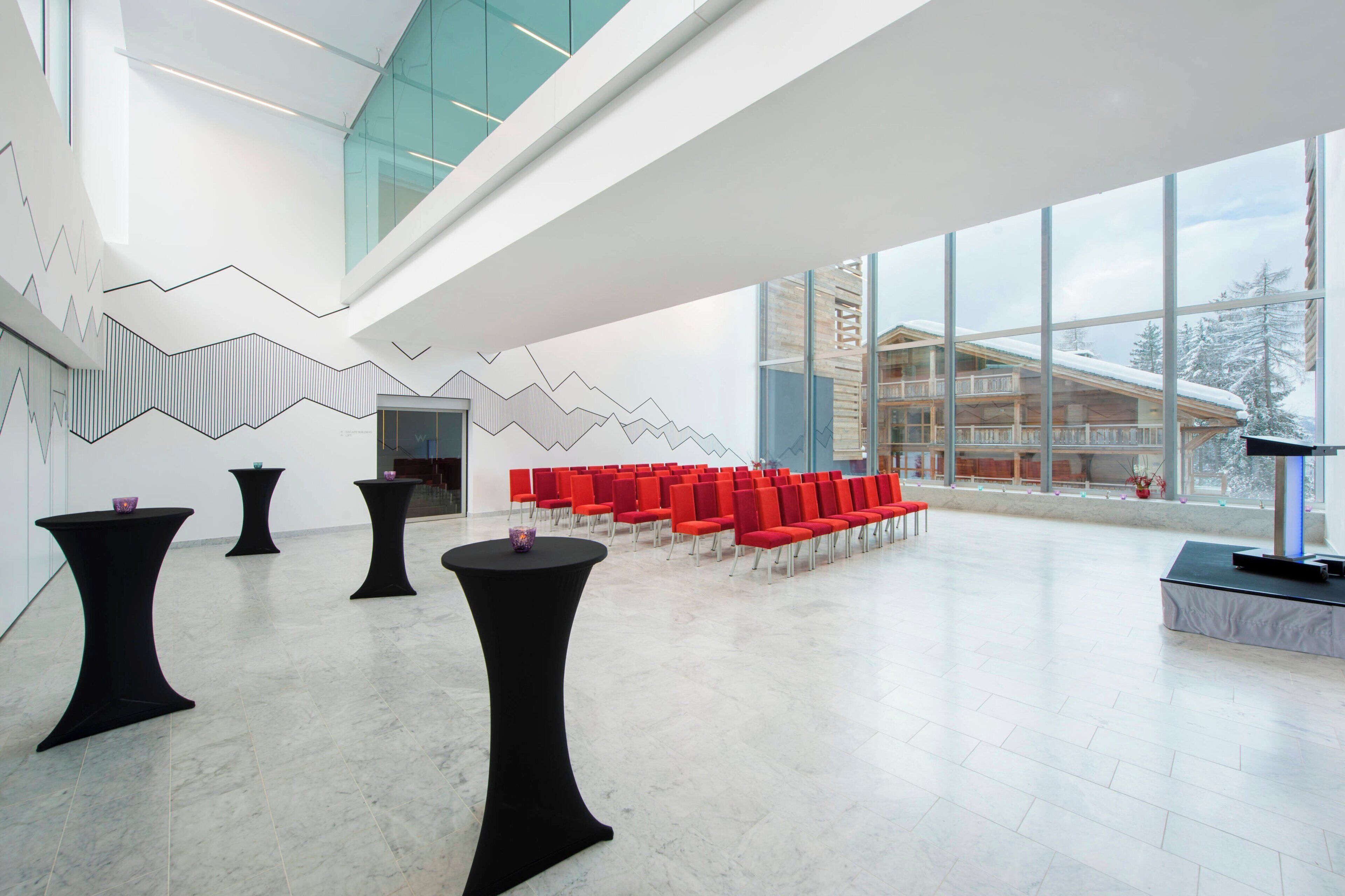 Un amplio salón con diseño moderno, que incluye una zona de estar con sillas rojas y un escenario con un atril.
