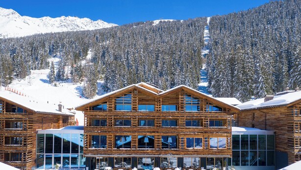 A multi-story wooden lodge with large windows, surrounded by snow-covered mountains and trees. - W Verbier (Bagnes)