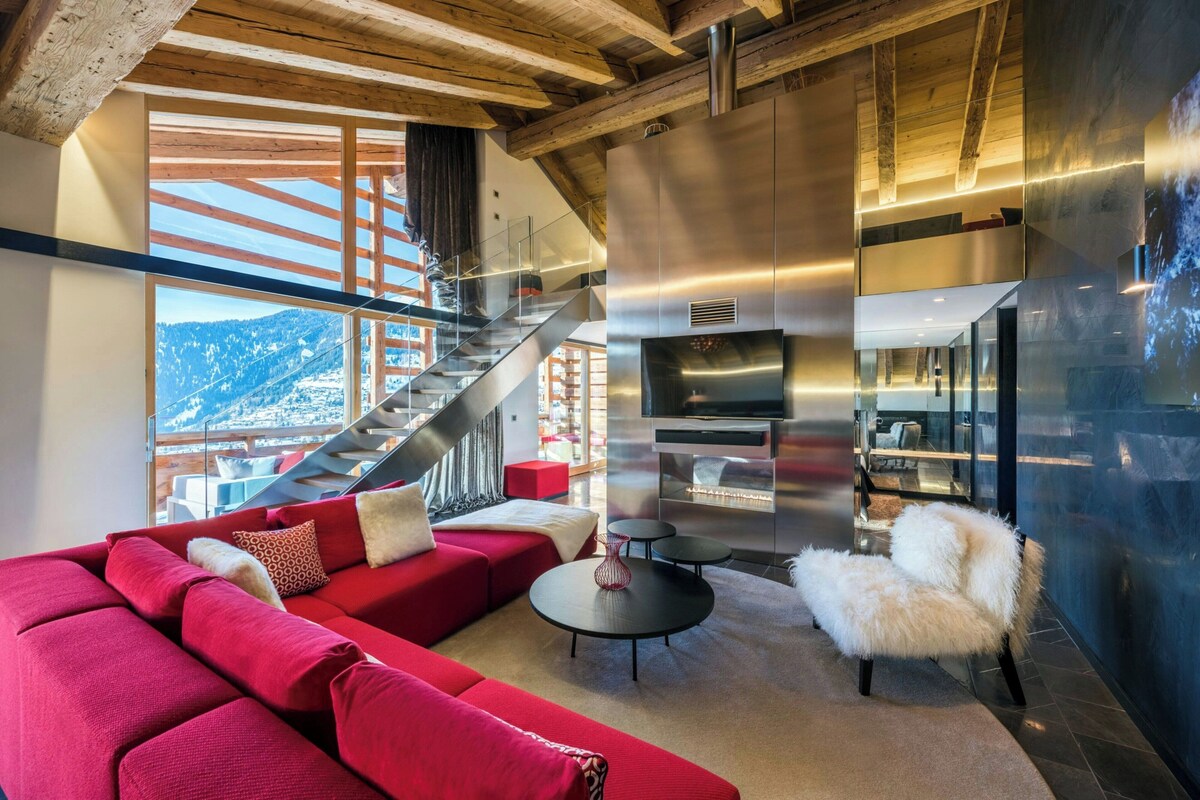 A modern living room with a red sofa, a white furry chair, a black coffee table, and a large TV. A staircase leads to a balcony with a view of a snowy landscape.