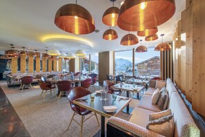 Un restaurante moderno con varias zonas de comedor, amplias ventanas con vistas a la montaña e iluminación colgante de cobre.