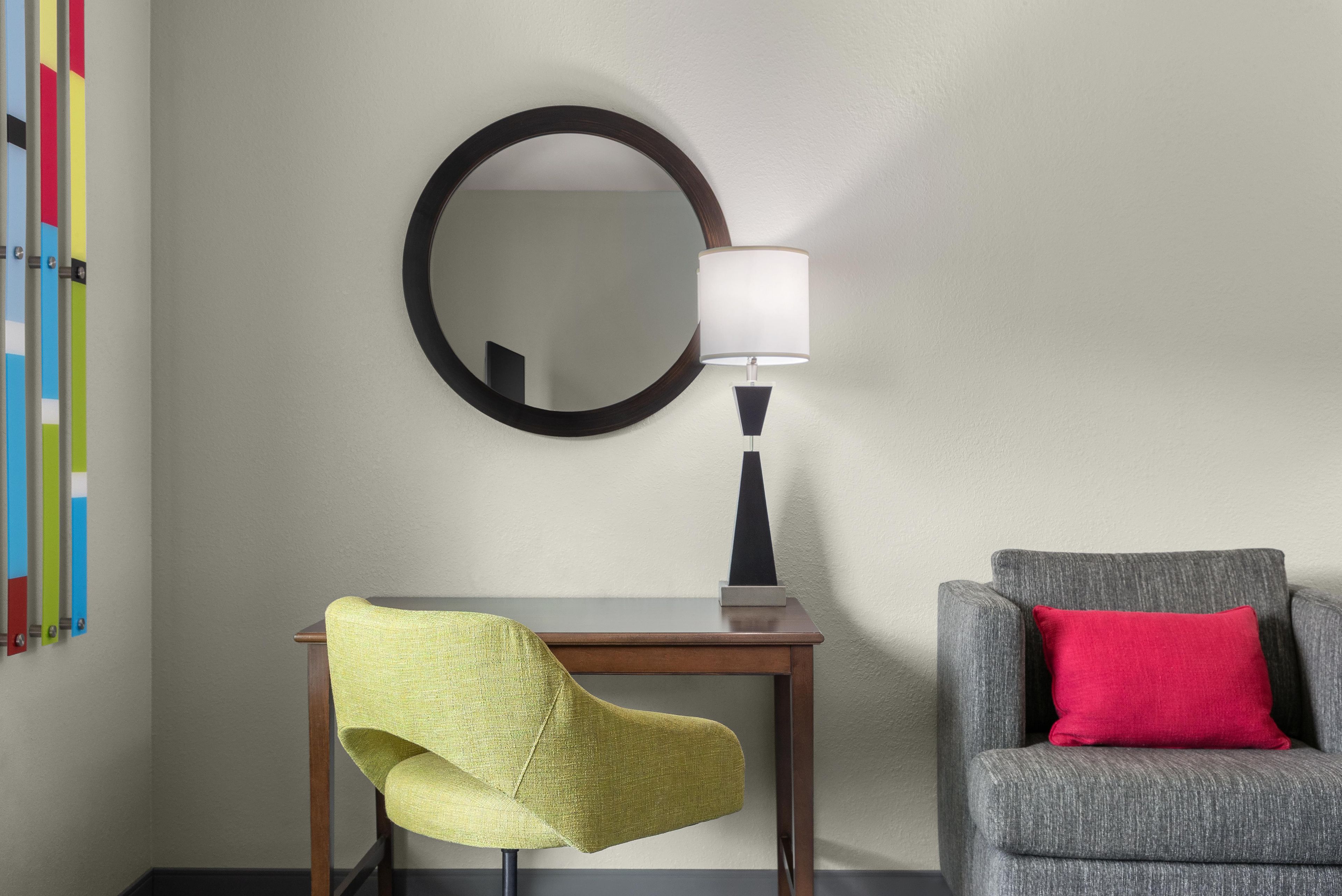 Una habitación con escritorio, una silla, una lámpara, un espejo y un sofá con un cojín.