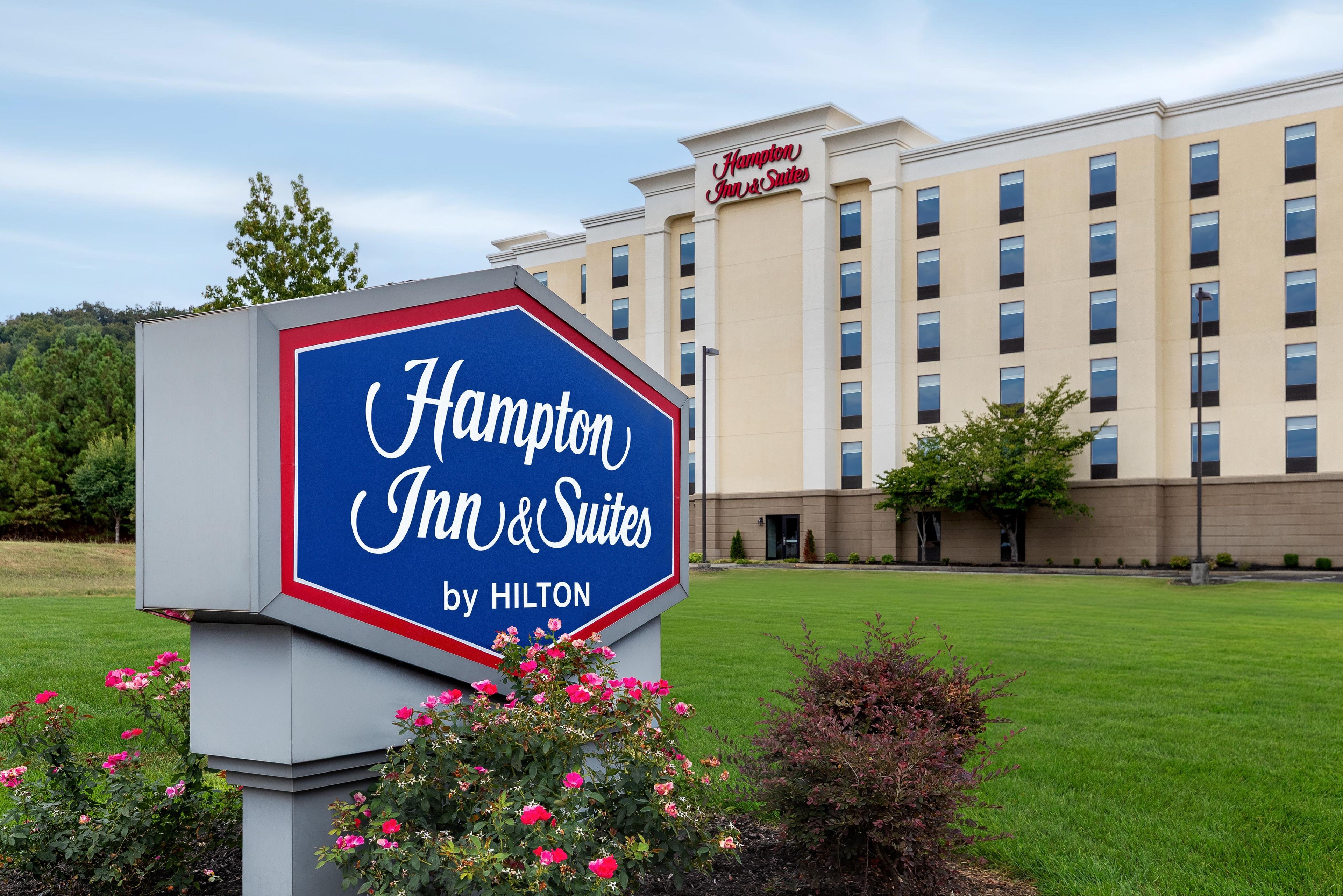 Un letrero de Hampton Inn & Suites con el logotipo de Hilton, rodeado de follaje y un edificio de hotel al fondo.