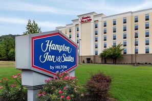 Skilt for et Hampton Inn & Suites hotel med Hilton-logo, omgivet af grøn bevoksning og en hotelbygning i baggrunden.