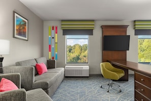 A hotel room with a gray sofa, a yellow chair, a desk, a TV, a window with a view of trees, and a colorful abstract art piece on the wall. - Hampton Inn & Suites Adairsville-Calhoun Area (Adairsville)