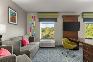 Quarto de hotel com sofá cinza, poltrona amarela, escrivaninha, televisão, janela com vista para árvores e uma obra de arte abstrata colorida na parede.