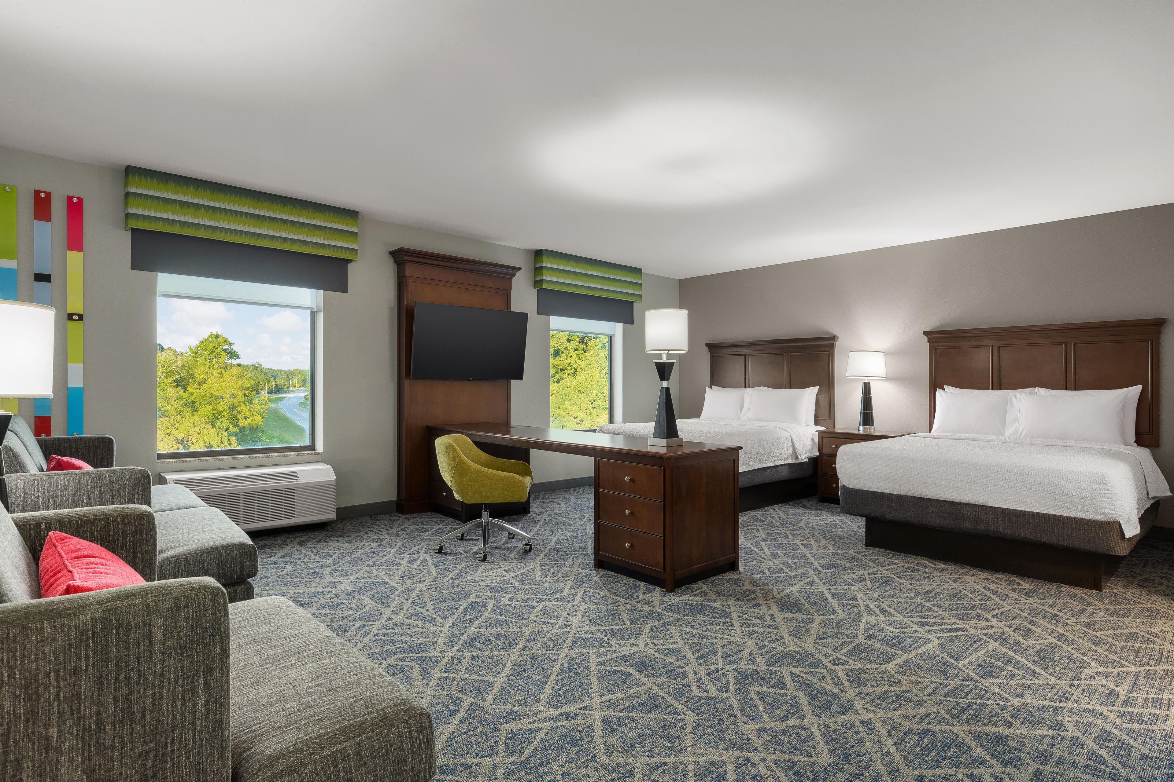 A hotel room with a bed, desk, chair, and a view of greenery outside.