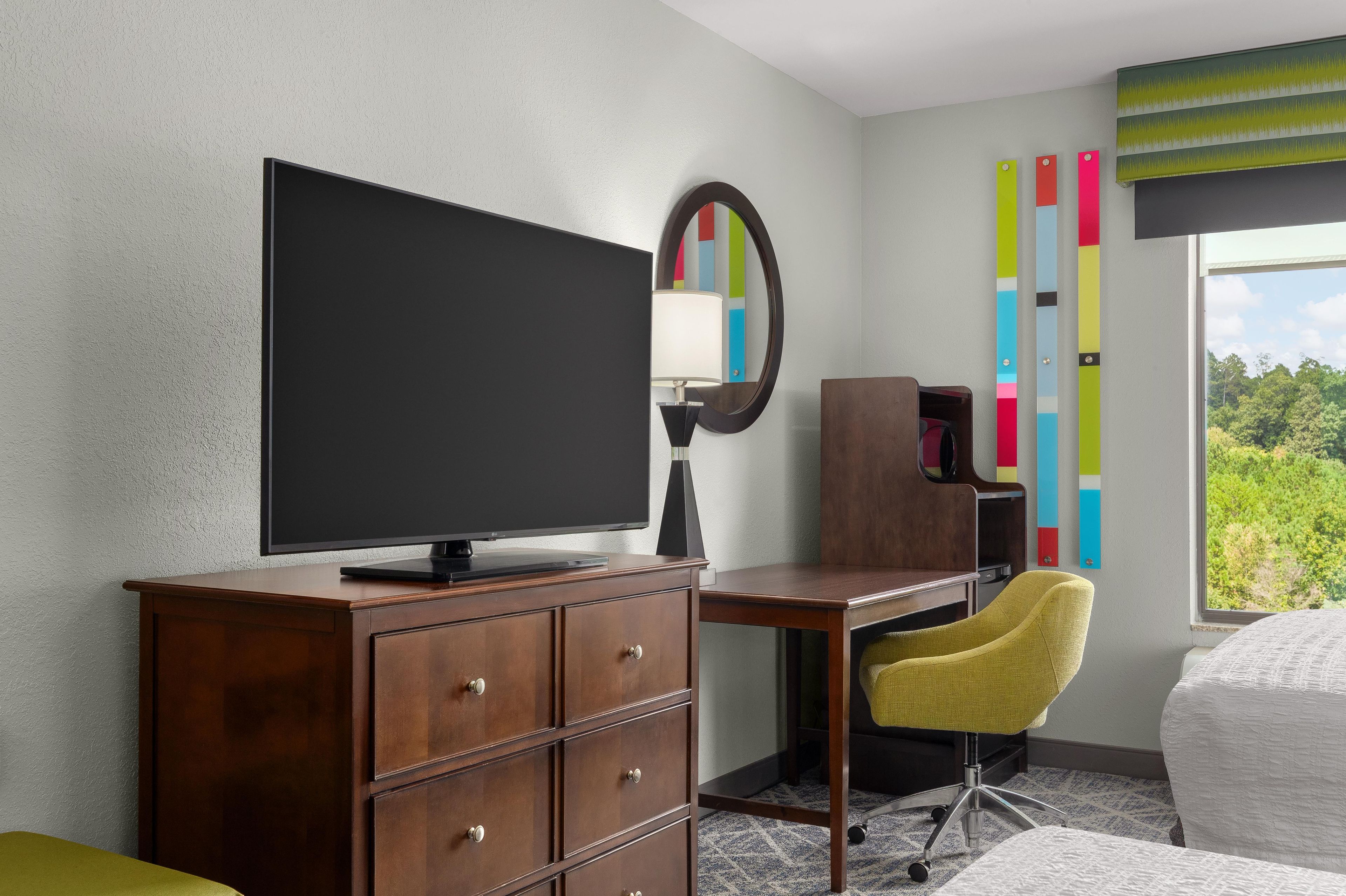 Une chambre d’hôtel avec un bureau en bois, une télévision, un miroir, une chaise jaune et un lit.