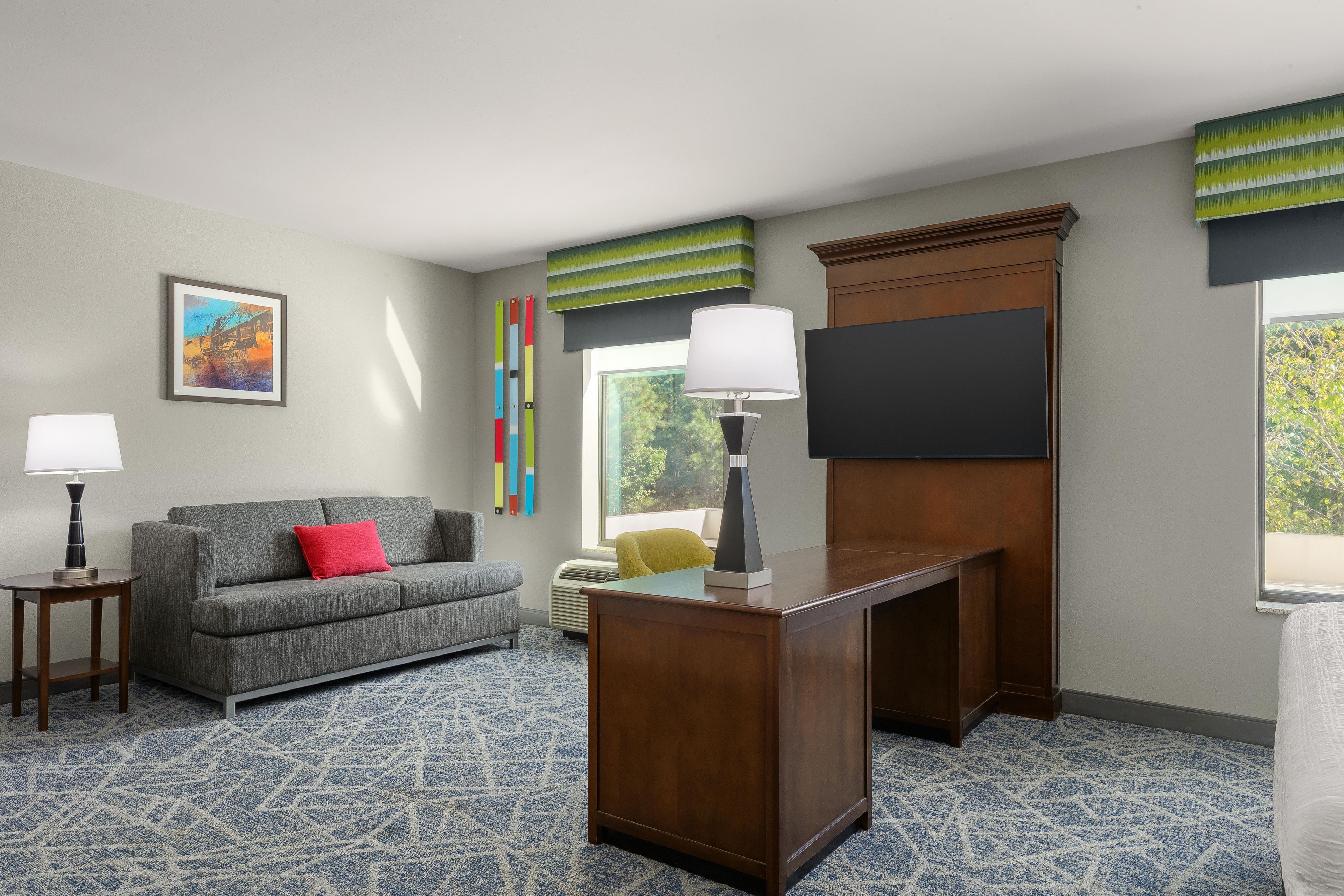 A hotel room with a gray sofa, a wooden desk, a TV, a framed picture on the wall, and a window with a view of trees.