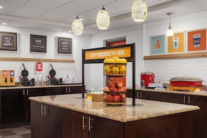 Estação de café da manhã com placa indicando o horário de início, variedade de frutas e máquina de café.