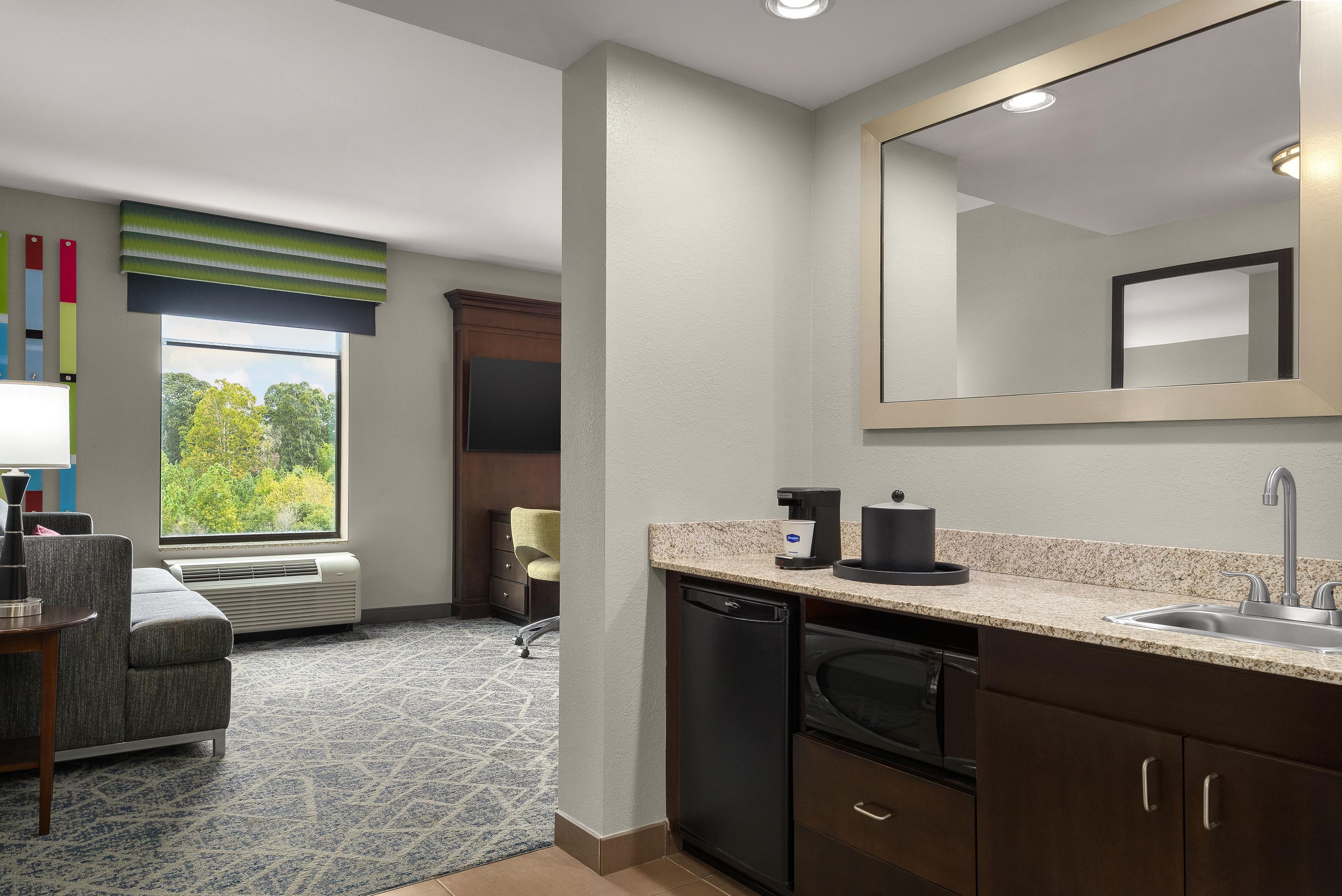 Habitación de hotel con kitchenette, un espejo grande, una ventana con vistas a árboles, un sofá y una silla.