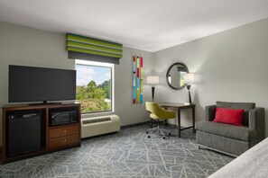 Een hotelkamer met een flatscreen televisie, een bureau met een lamp, een stoel, een bank met een rood kussen en een raam met uitzicht op groen.