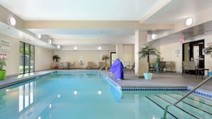 Una piscina interior amb aigua cristal·lina, envoltada de gandules i taules, i equipada amb senyals de seguretat i indicadors de sortida d'emergència.