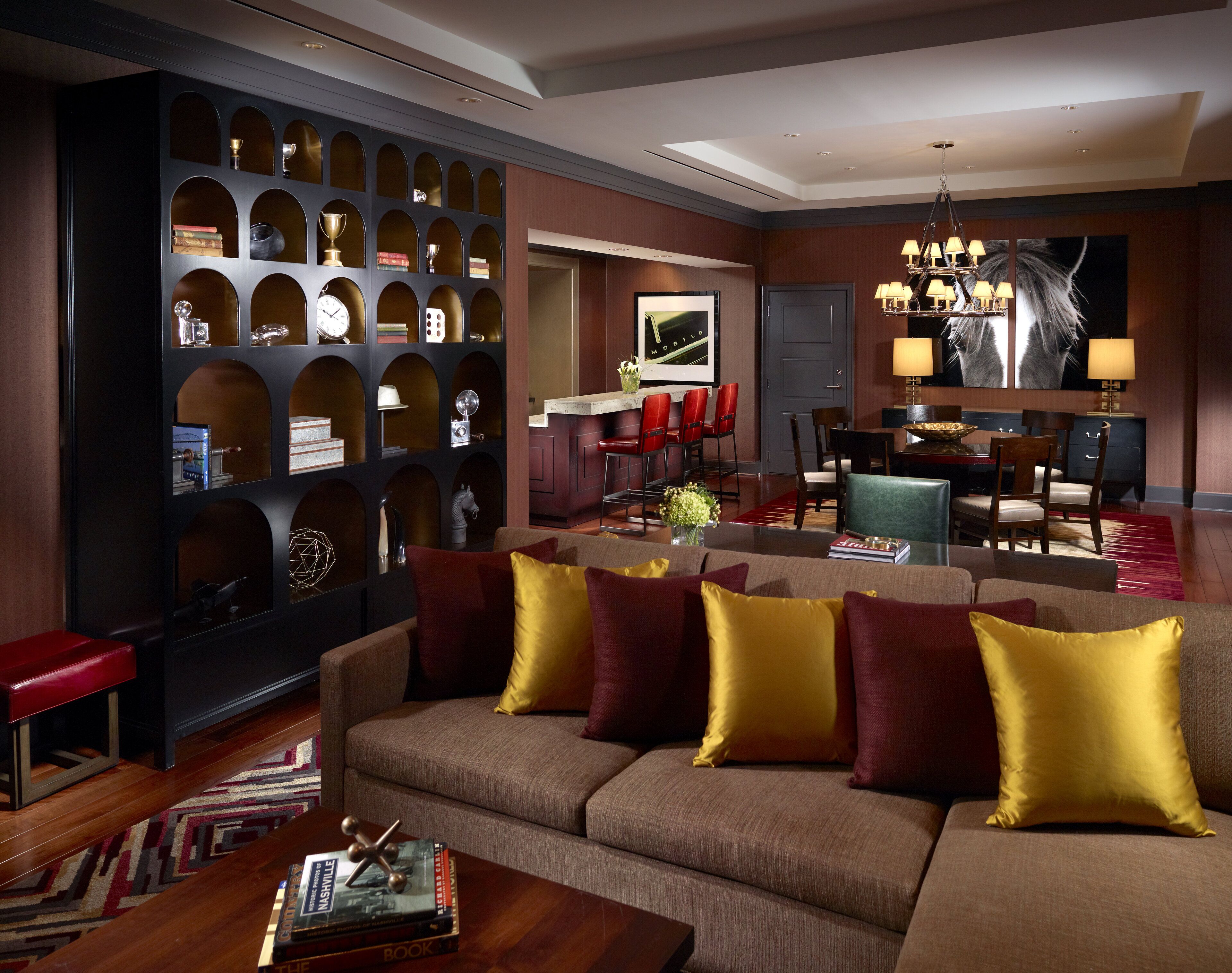 A modern living room with a sectional sofa, a coffee table with books, a wall-mounted shelf with decorative items, a dining area with red chairs, and a chandelier.