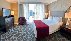 A hotel room with a large bed, a TV on a dresser, a chair, and a view of the city through the window.