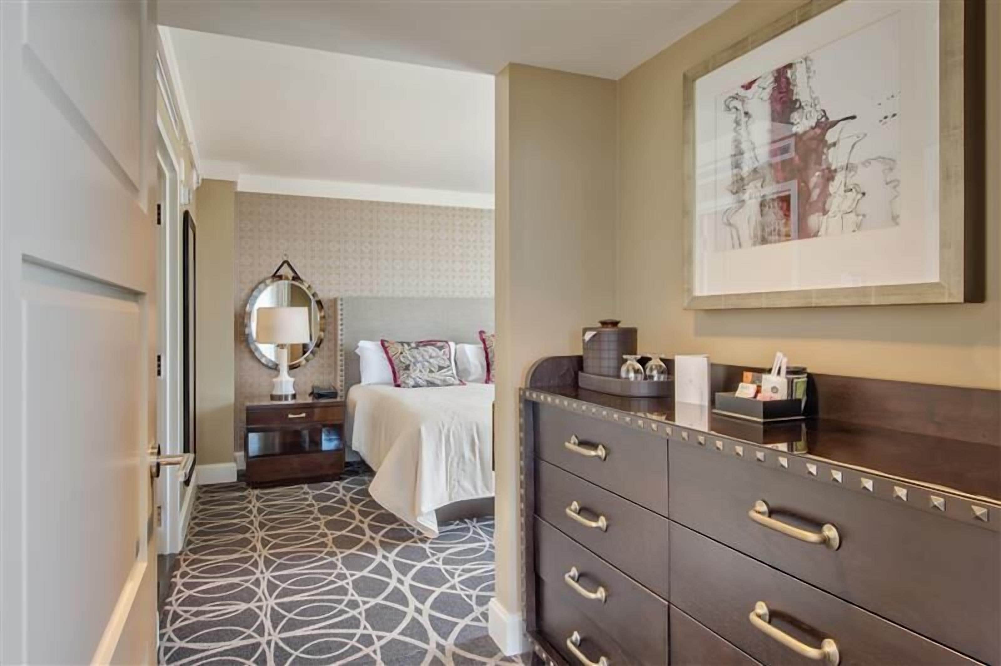 Quarto de hotel com piso estampado, cama com roupa de cama branca, cômoda com gavetas, espelho e uma obra de arte emoldurada na parede.