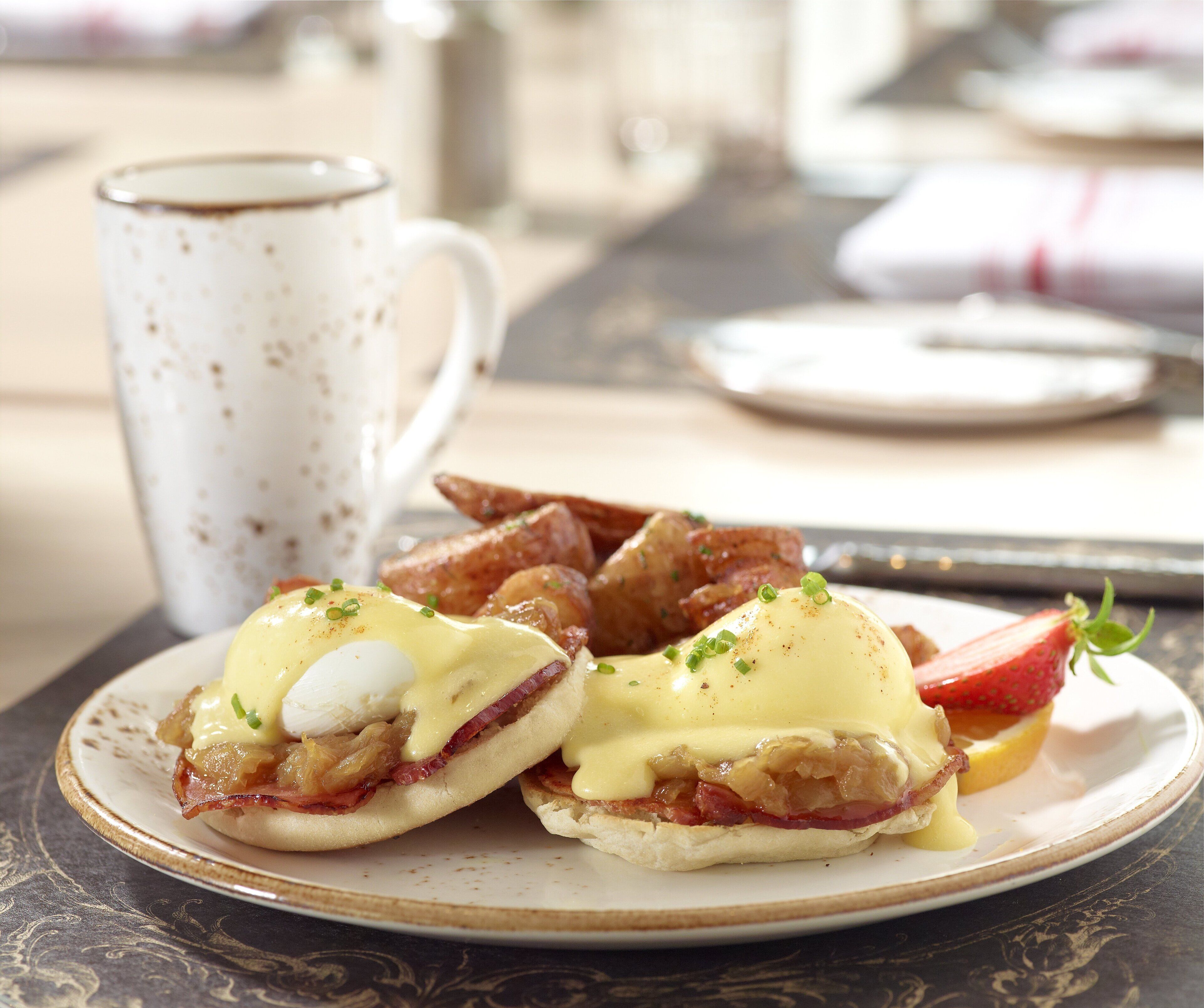 A plate of eggs Benedict with bacon and hollandaise sauce, accompanied by a cup of coffee and croissants.