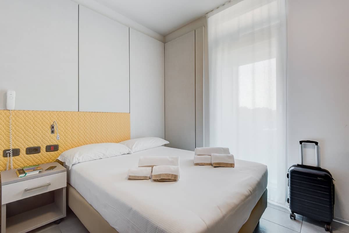 A hotel room with a bed, a nightstand, a suitcase, and a window with curtains.