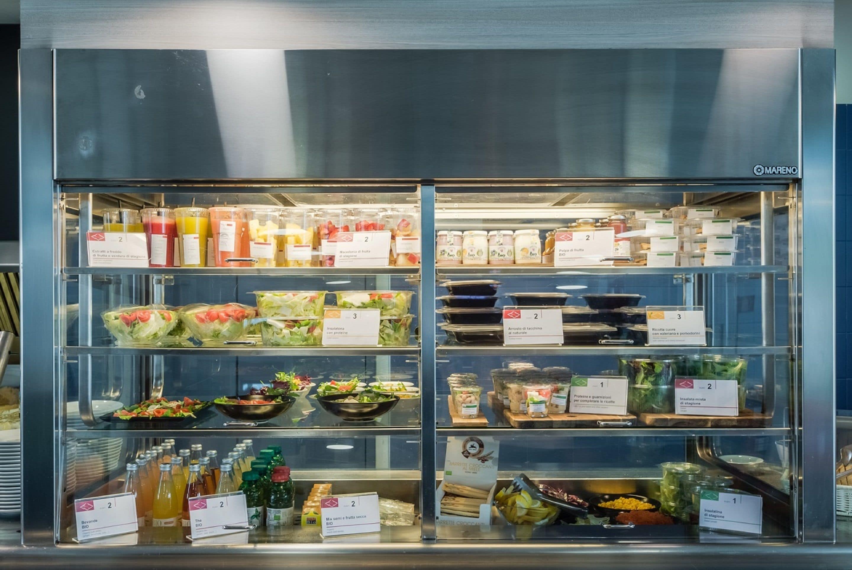 Une vitrine réfrigérée contenant divers produits alimentaires et condiments, notamment des salades, des sandwichs et des boissons.