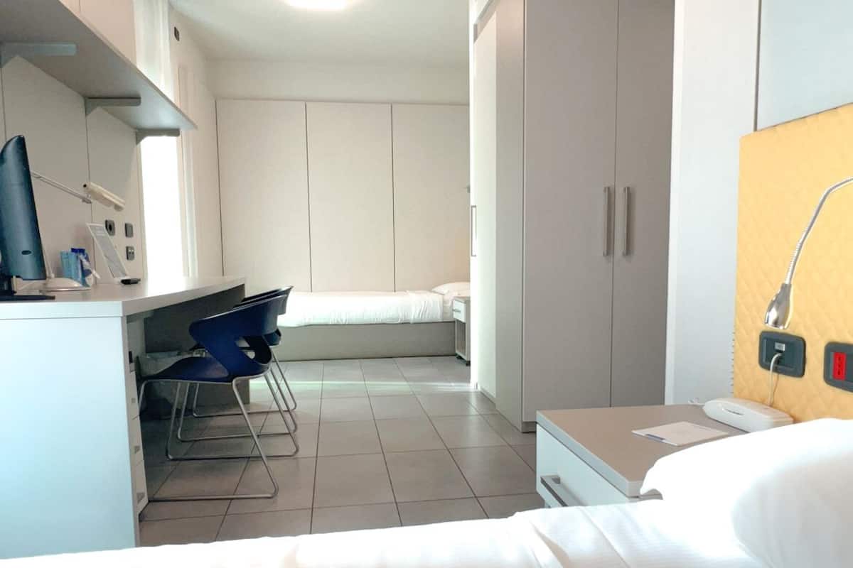A hotel room with a bed, a desk with a chair, a wardrobe, and a bedside table with a lamp.