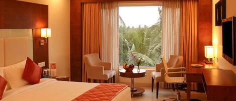 A hotel room with a large bed, two armchairs, a desk, and a view of greenery through the window.