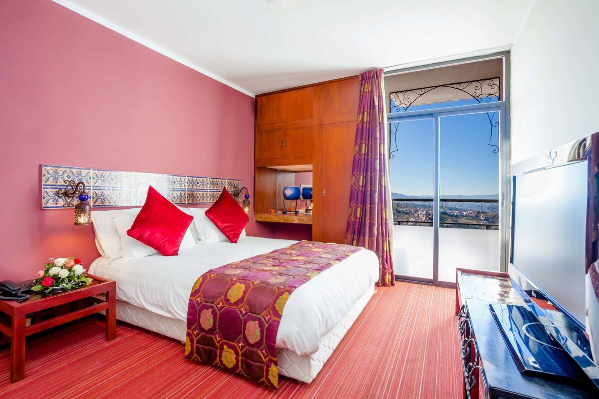 Habitación de hotel con cama, mesita de noche, escritorio, ventana con vistas y balcón.