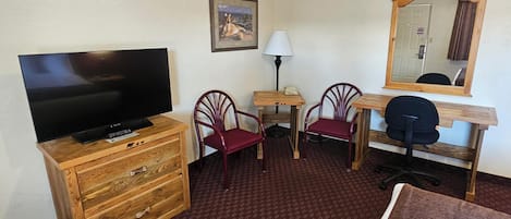 Hotelkamer met een houten dressoir, een televisie, twee stoelen, een bureau met een lamp en een grote spiegel.