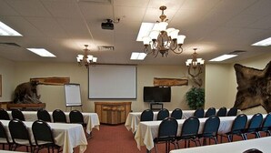Un salón de conferencias con mesas y sillas, un proyector, una pantalla, un televisor y elementos decorativos como una escultura de oso y una gran piel de animal.