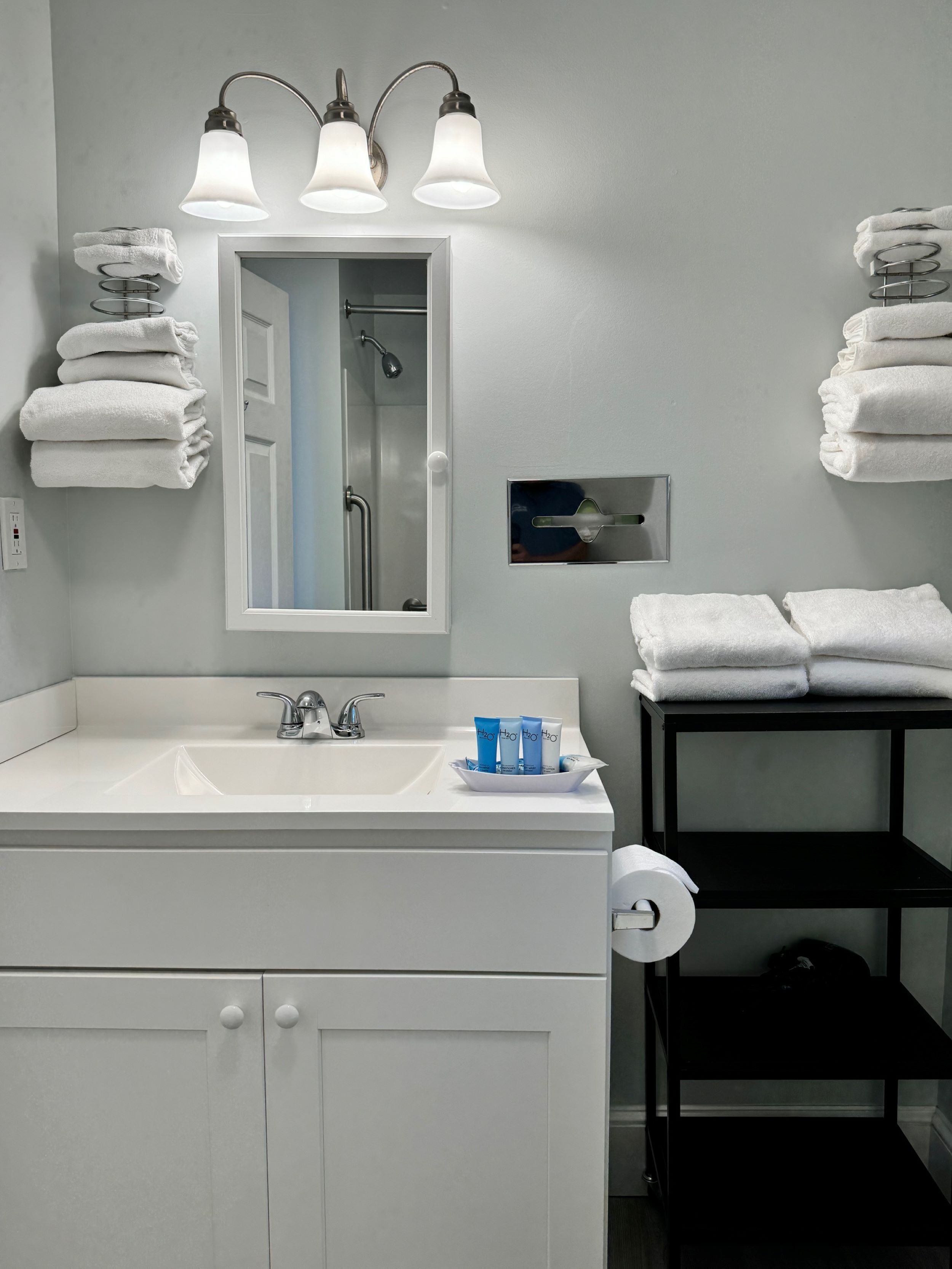 Une salle de bain moderne avec un lavabo blanc, un miroir et trois appliques murales. Des serviettes sont pliées sur un porte-serviettes et une étagère noire contient d’autres serviettes.