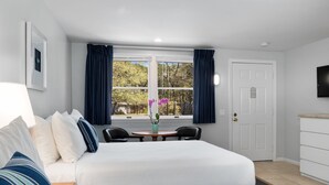 A hotel room with a bed, a desk, a chair, a TV, and a window with curtains. - The Even'tide (Wellfleet)