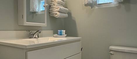 A bathroom with a white vanity, a sink, a mirror, a toilet, and a window with curtains.