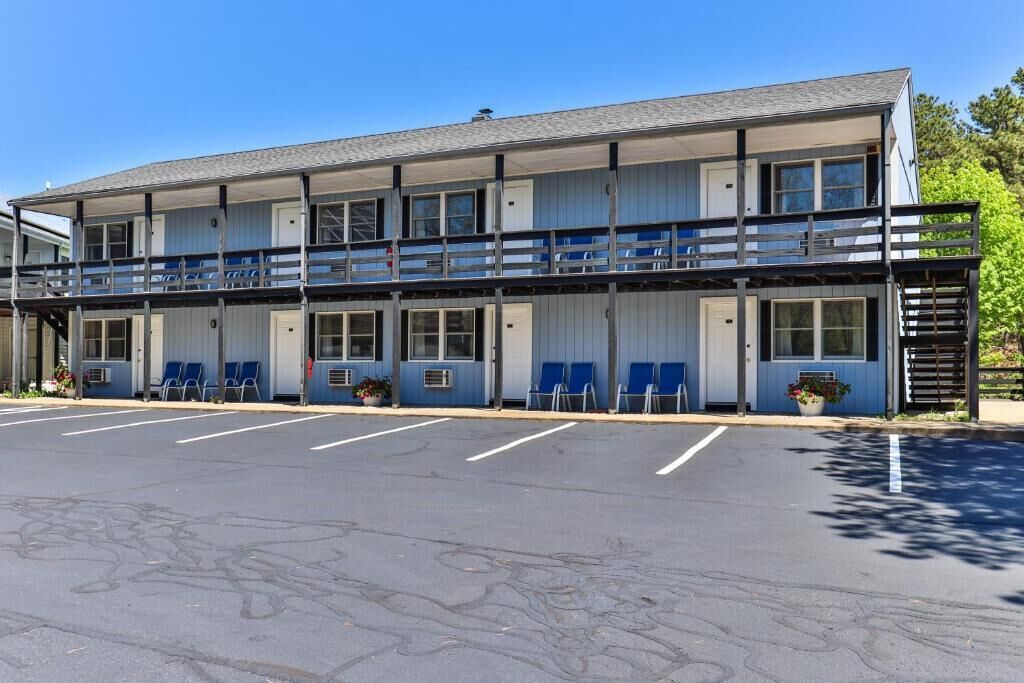 A two-story motel with a balcony on the upper floor, a parking lot in front, and a row of blue chairs outside.