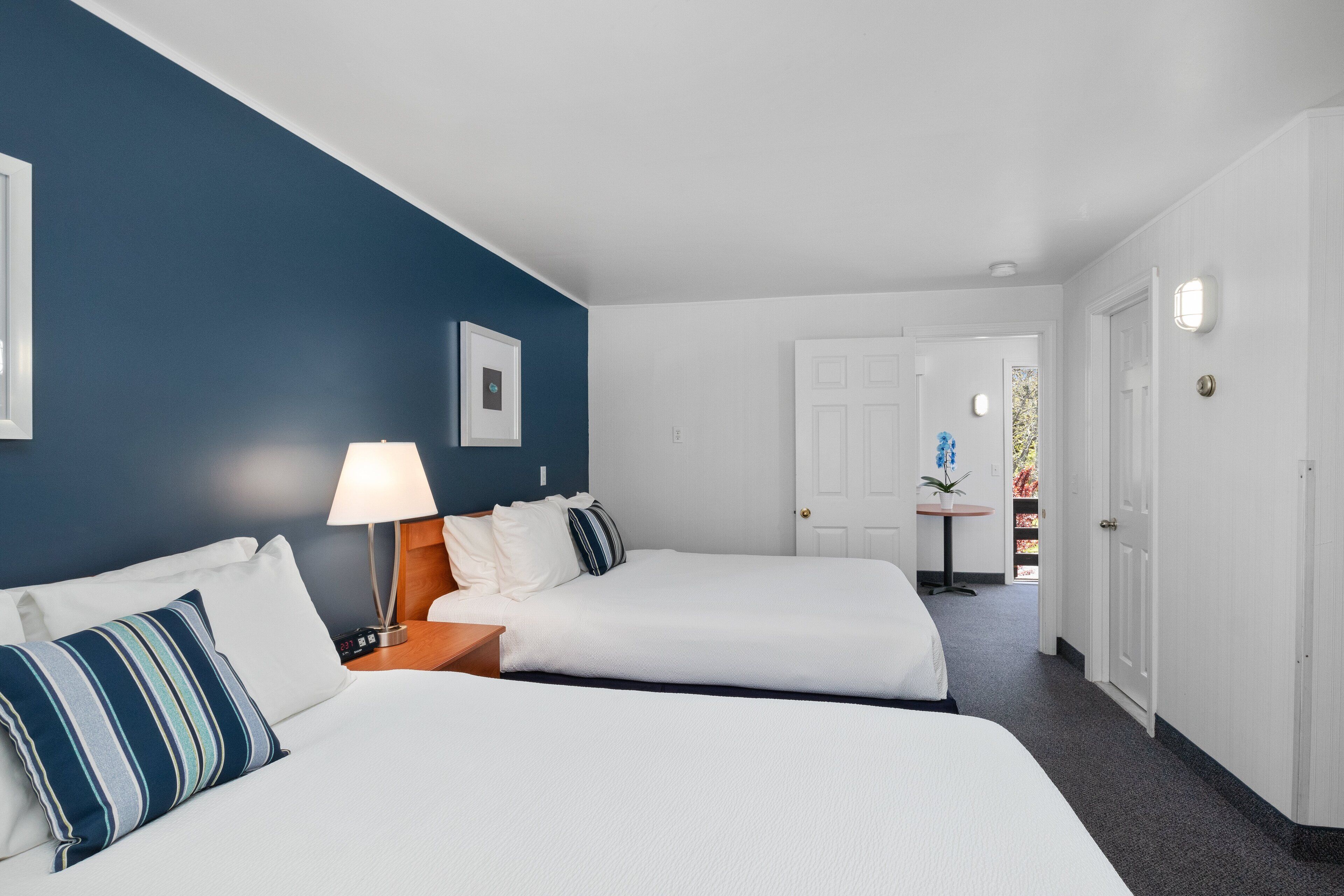 Ein Hotelzimmer mit zwei Betten, einem Nachttisch mit Lampe, einem gerahmten Bild an der Wand und einer Tür zu einem weiteren Raum.