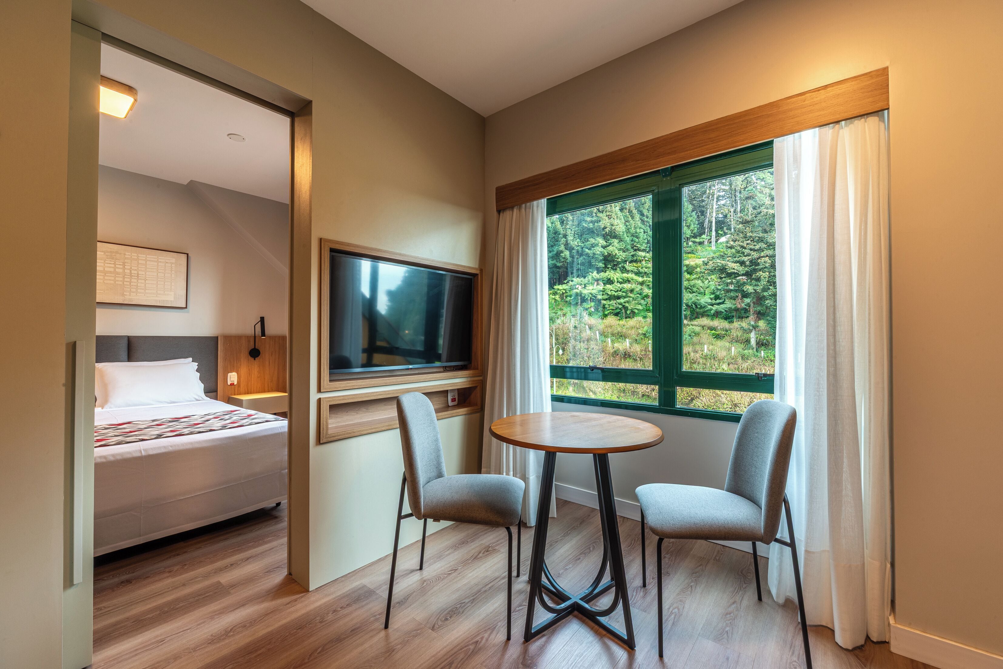 Une chambre d’hôtel avec un lit, une télévision, une petite table, deux chaises et une fenêtre donnant sur la forêt.