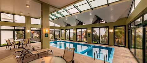 A spacious indoor swimming pool area with clear glass walls, a lounge with chairs and tables, and a view of the surrounding landscape.
