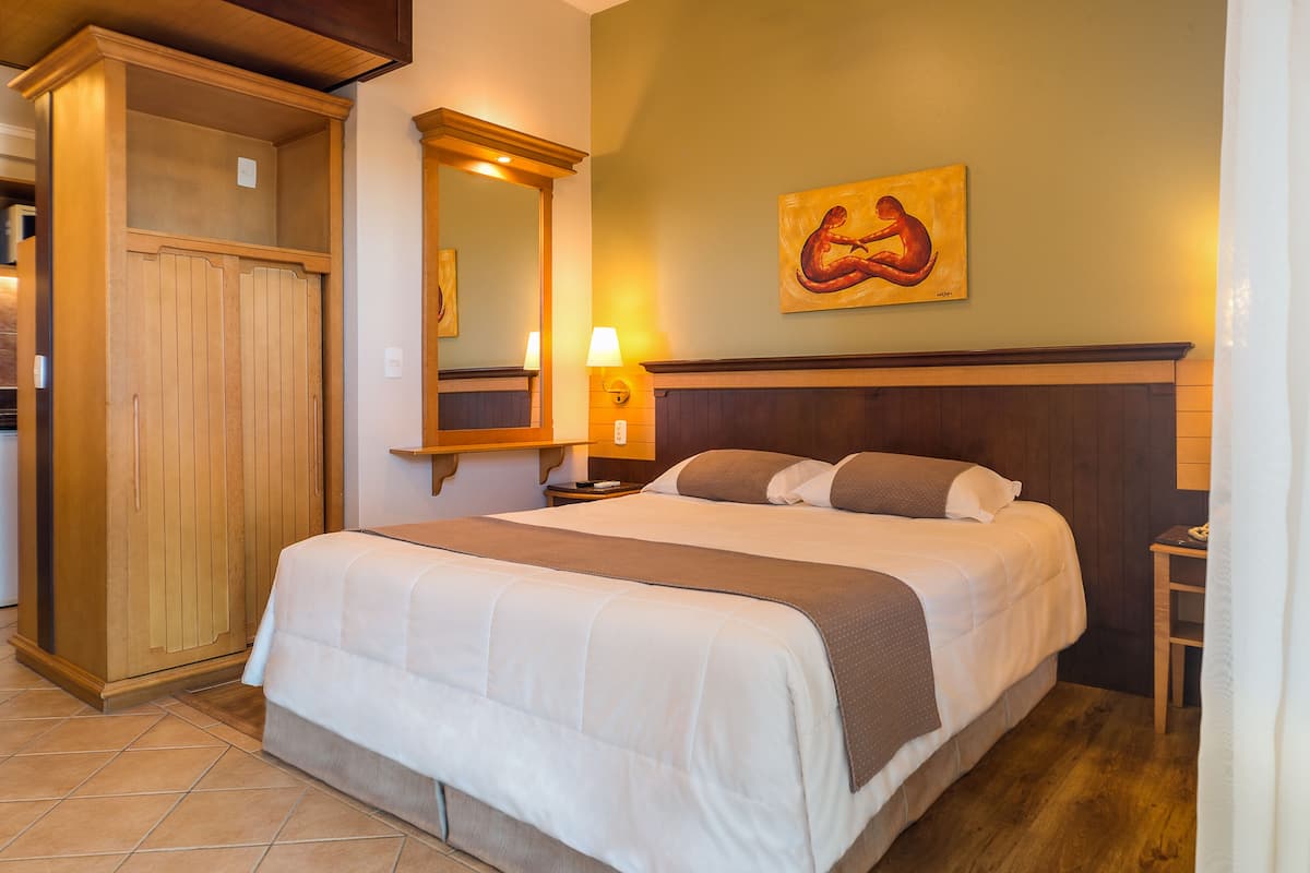 Habitación de hotel con una cama grande, un armario de madera, un cuadro en la pared y una mesita de noche con lámpara.