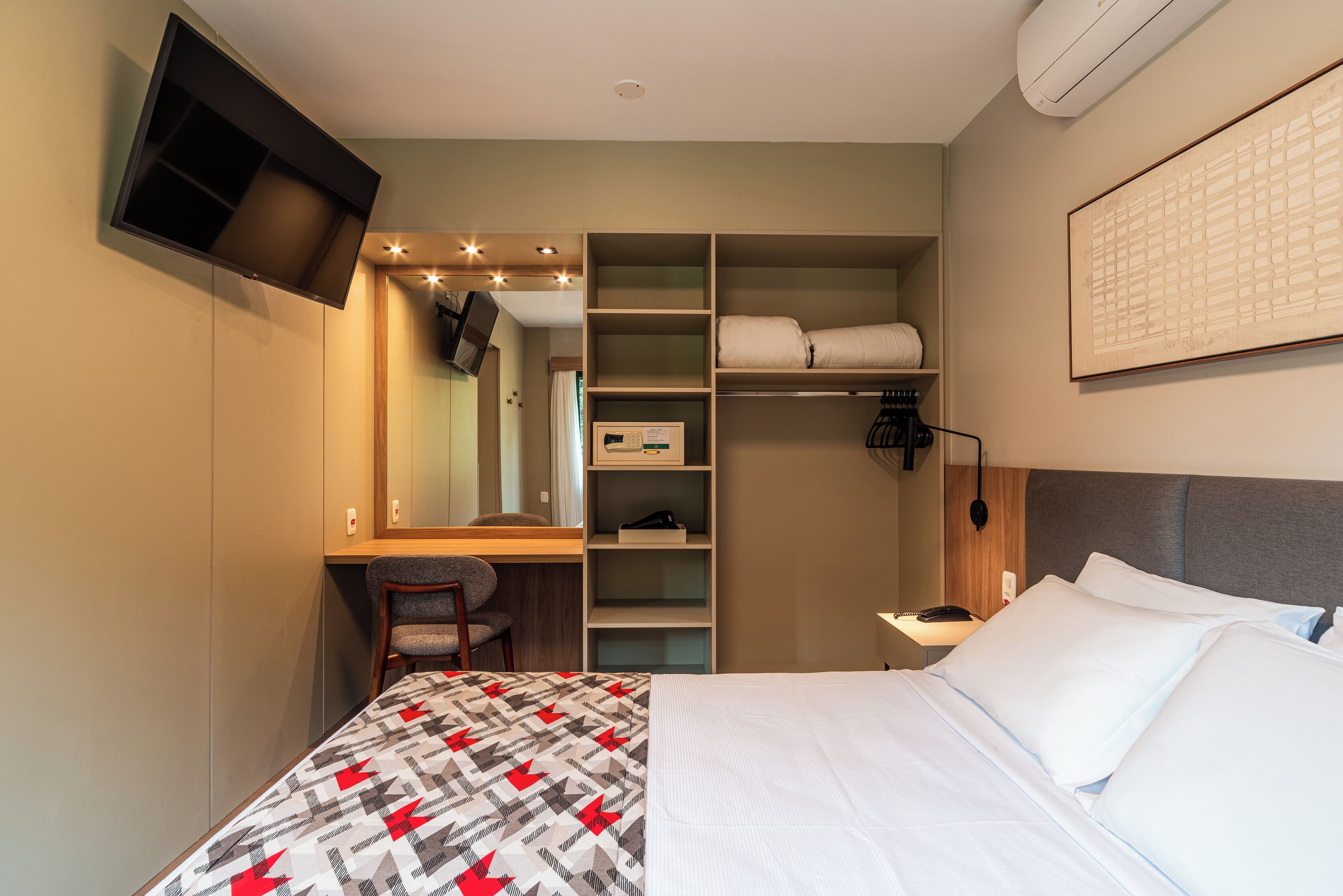 Habitació d'hotel amb un llit, un escriptori amb cadira, un armari, una taula de nit i una televisió penjada.