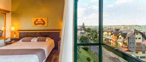 A hotel room with a large window, a bed, a ceiling fan, and a painting on the wall.