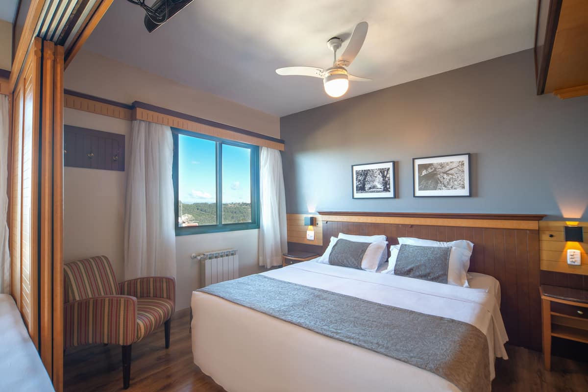 Una habitación de hotel con una cama grande, una silla, una ventana con cortinas, un ventilador de techo y dos cuadros en la pared.