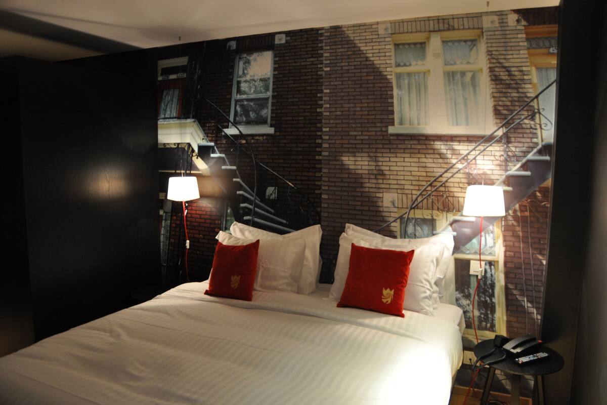 A hotel room with a bed, two red pillows, a nightstand, a lamp, and a view of a brick building with a window and a staircase.