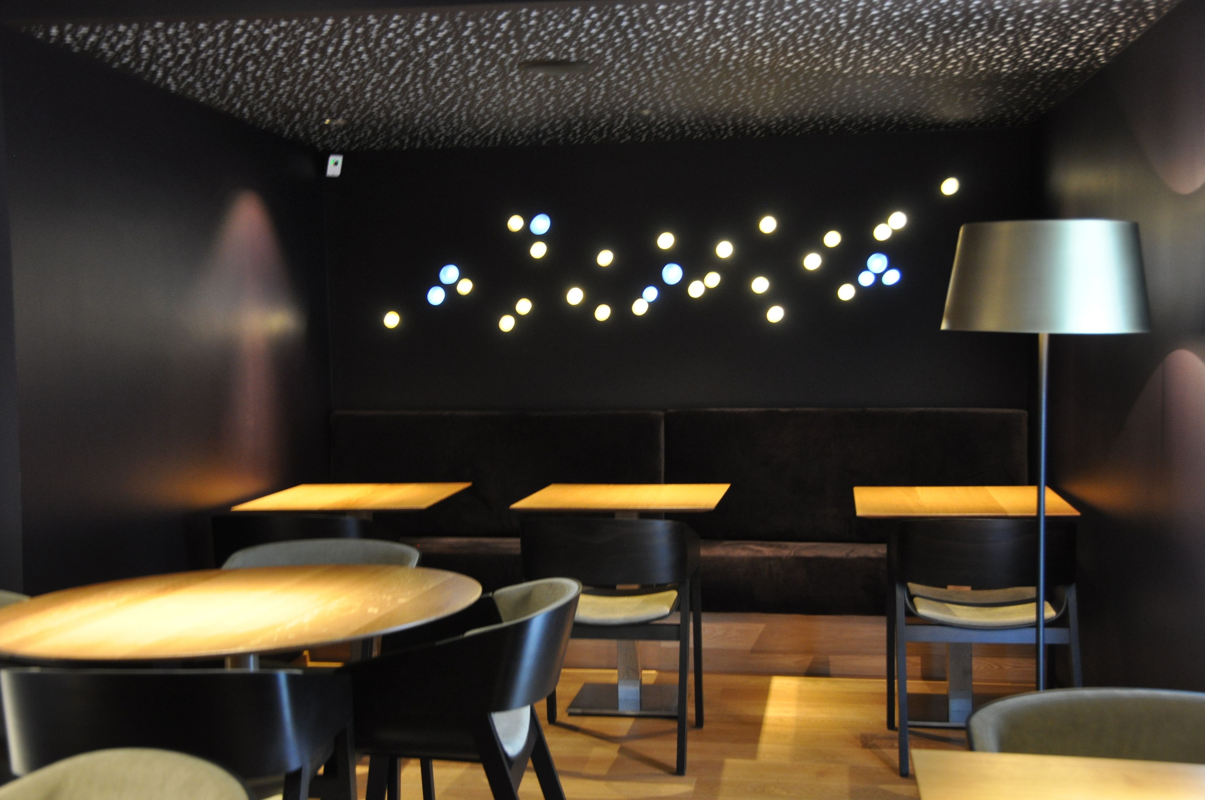 Un intérieur moderne avec des tables et des chaises rondes, un mur noir orné d’une guirlande lumineuse et un lampadaire.